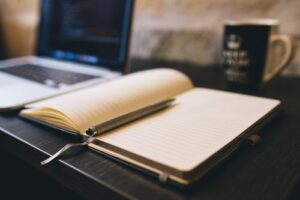 A copywriting company home set up. with notebook in the foreground and laptop and mug behind.